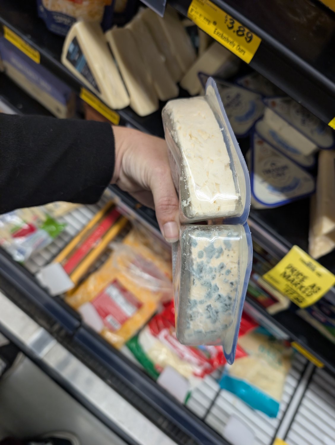 A hand holding two packages of blue cheese in front of a grocery store shelf, one cheese has no visible spots of blue mold, while the other is covered in spots.